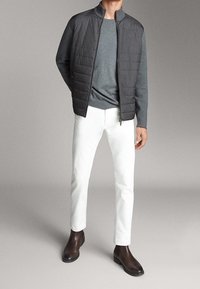 Giubbotto grigio trapuntato con colletto alto, indossato sopra un maglione grigio scuro e pantaloni bianchi slim-fit, abbinato a stivali alla caviglia marroni.