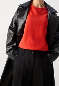 Manteau en cuir noir sur un pull rouge, présentant un tissu texturé. Pantalon noir avec un détail de bouton unique et des plis.