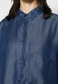 Femme portant une chemise en denim bleu foncé délavée avec boutons, col et poche poitrine, visible du cou jusqu'au milieu du torse.