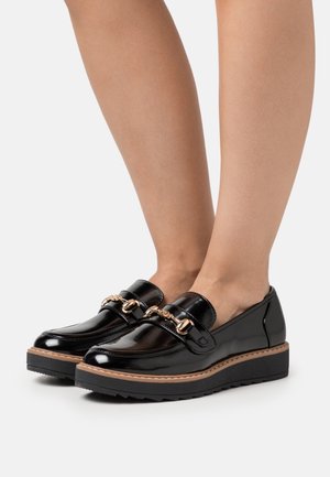 Des pieds portant des mocassins noirs brillants avec détail en chaîne dorée et semelles épaisses en noir, se tenant sur une surface blanche.