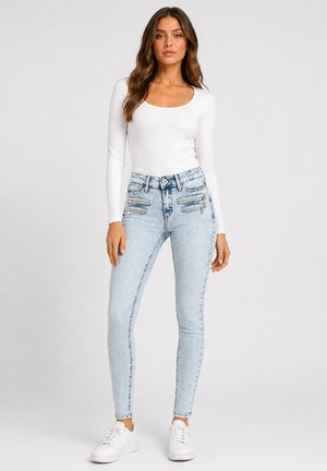 Kvinde stående iført hvid langærmet top, lyseblå skinny jeans med lynlåse foran og hvide sneakers mod en ensfarvet baggrund.