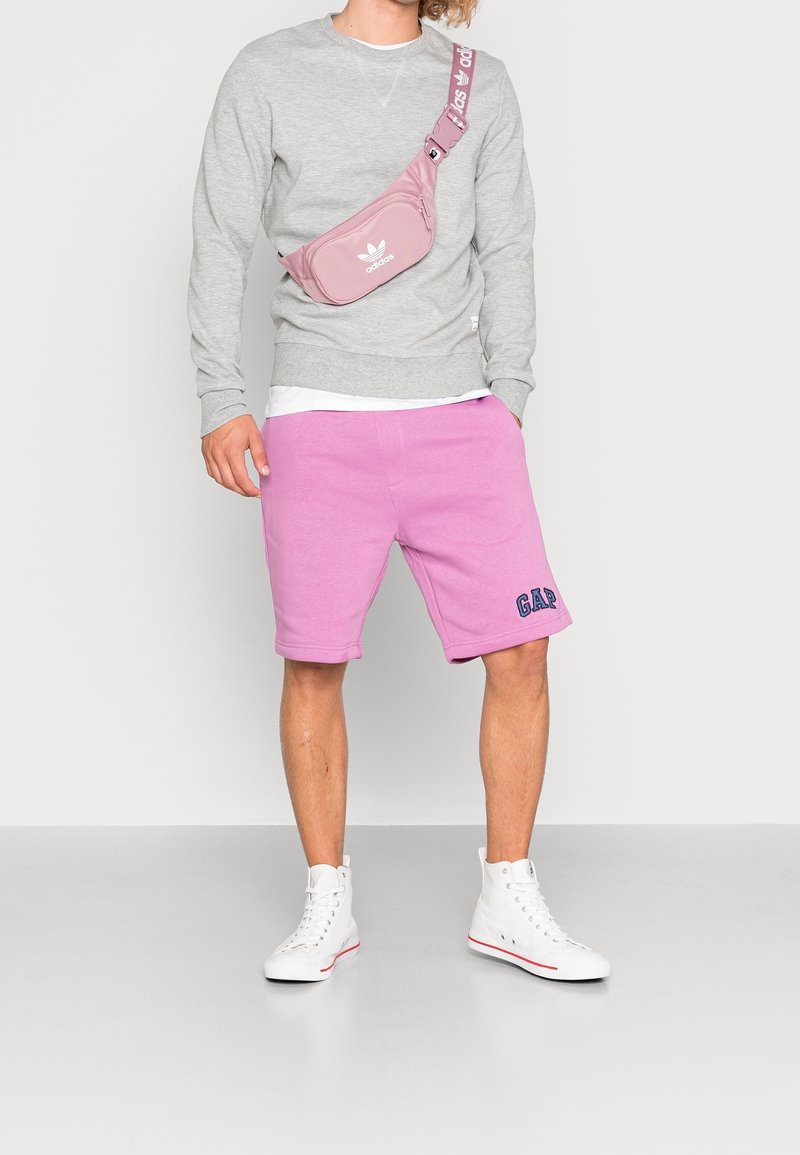 Sudadera gris, pantalones cortos rosas con el logo de GAP y zapatillas altas blancas. La bolsa es rosa con el logo y la correa blanca de Adidas.