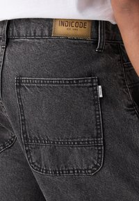 Mörkgrå denimjeans med en enda bakficka, med en brun läderlapp märkt "INDICODE EST. 1993" vid midjan.