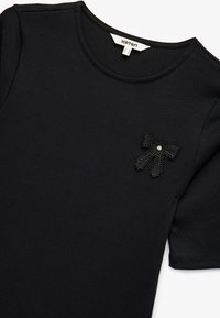 Schwarzes geripptes T-Shirt mit kurzen Ärmeln, das an der oberen linken Ecke mit einer Spitzen-Schleifenakzent und einem Strassstein verziert ist.