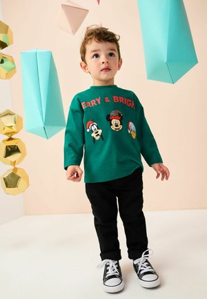 Grøn langærmet skjorte med tegneseriefigurer og teksten "Merry & Bright". Matcher med sorte bukser og sorte sneakers med hvide snørebånd.