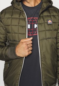 Man in een olijfgroene gewatteerde jas met ellesse-logo, gedeeltelijk open geritst om een zwart graphic t-shirt met een rood wereldontwerp te onthullen.