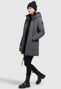 Harmaa puffcoat, jossa on chevron-ommeltu pinta, vetoketjukinnitys, säädettävä huppu ja sivutaskut. Yhdistetty mustiin legginsseihin ja paksuihin mustiin saappaisiin.