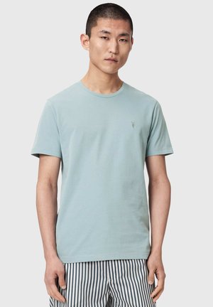 Nuori mies, joka pukeutunut vaaleansiniseen lyhythihainen t-paitaan ja pystysuoraan raidoitettuihin mustavalkoisiin housuihin, seisoo yksivärisen taustan edessä.