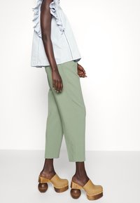 Lichtblauw ruffles-top met een losse fit, gecombineerd met groene taps toelopende broek en bruine houten plateauklompen met ronde zolen.
