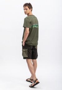 Olijfgroene katoenen t-shirt met een grafisch ontwerp op de achterkant, gecombineerd met zwarte cargoshorts met zijzakken en contrasterende accenten.