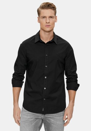 Camisa negra de manga larga con botones y cuello, que presenta un logotipo sutil y una suave tela de algodón. Llevada con jeans grises.