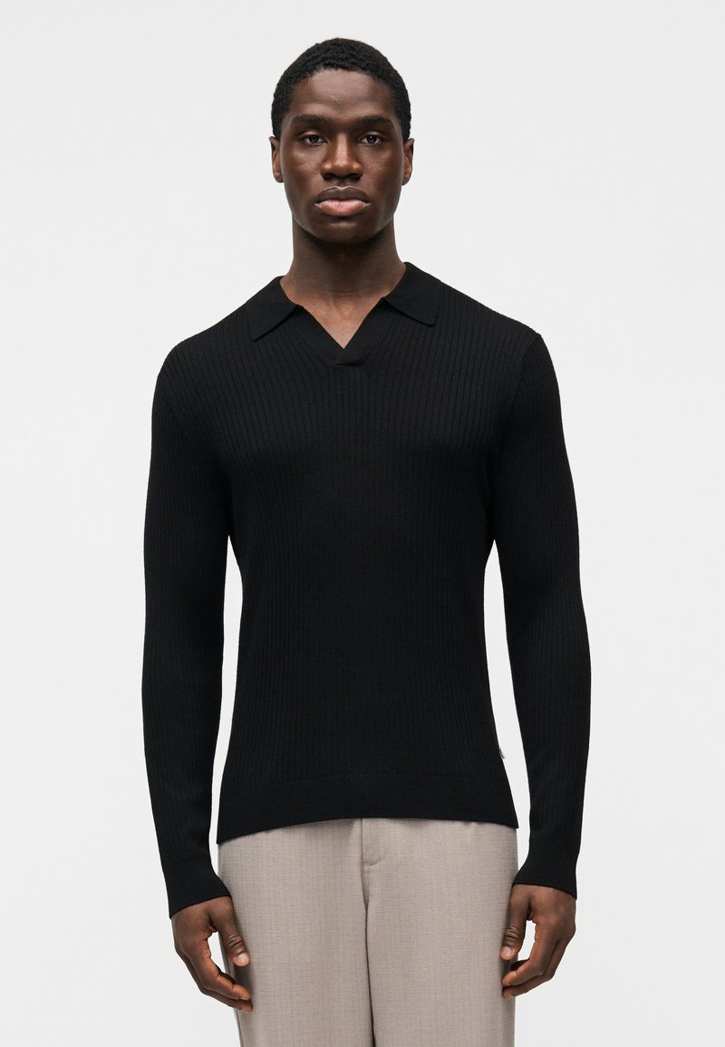 Pull noir côtelé à col en V et col court, doté de manches longues et d'une coupe ajustée, associé à un pantalon gris clair.