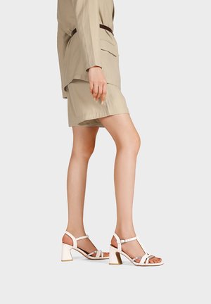 Donna che indossa un completo corto beige e sandali bianchi con tacco largo e cinturini, in piedi di lato contro uno sfondo uniforme.