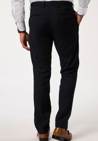 Pantalones navy con un corte ajustado presentan dos bolsillos traseros con cierres de botón, combinados con zapatos de cuero marrón, mostrados desde la parte posterior.