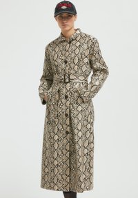 Dolga plašč v oblikovanju kačjega vzorca, z bež in črno barvno shemo, velikimi gumbi in stranskimi žepi. Teksturovan material.