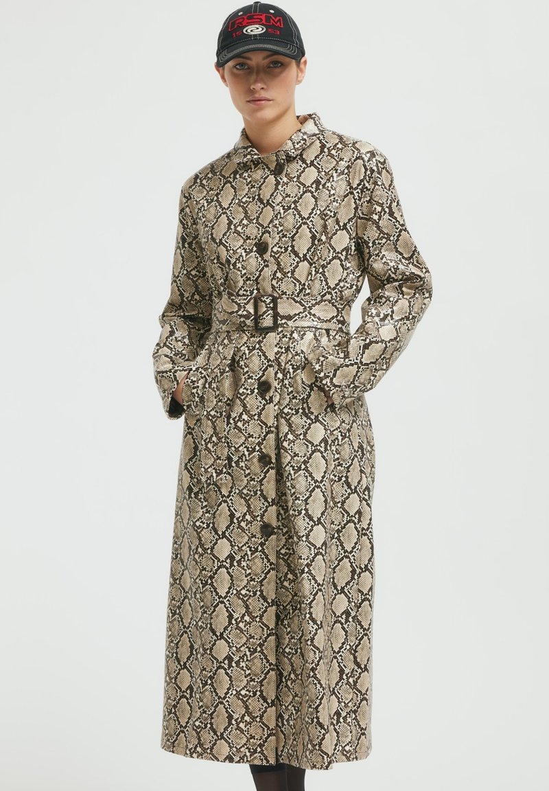 Dolga plašč v oblikovanju kačjega vzorca, z bež in črno barvno shemo, velikimi gumbi in stranskimi žepi. Teksturovan material.