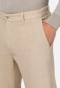 Pantaloni beige realizzati in tessuto liscio con vita aderente, dotati di tasche laterali in diagonale e chiusura a bottone. Dettaglio di texture sottile visibile.