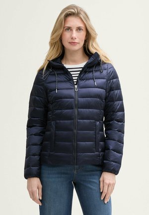 Veste matelassée navy avec capuche, fermeture éclair sur le devant et cordons élastiques. Présente un matelassage horizontal et deux poches avant. Associée à un haut rayé.