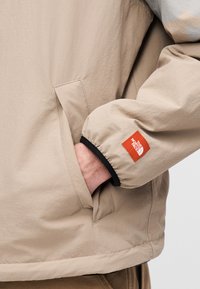 Μπεζ αντιανεμικό με ομαλή υφή, με μαύρο μανσέτι και εμφανές πορτοκαλί λογότυπο. Ορατή λεπτομέρεια πλαϊνής τσέπης.