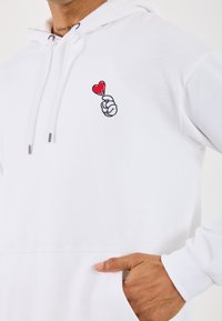 Felpa bianca in cotone, con un piccolo cuore rosso tenuto in mano tramite ricamo nero sul petto sinistro, dotata di tasca frontale.