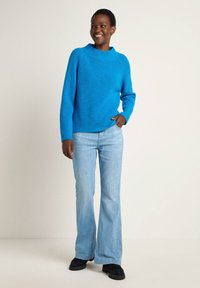 Helle blaue Strickjacke mit geripptem Muster, ausgestattet mit einem hohen Kragen und einer lässigen Passform, kombiniert mit hellblauen Schlagjeans und schwarzen Schuhen.