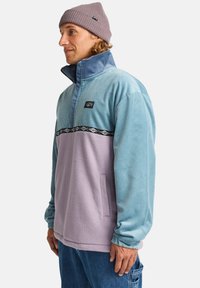 Pullover a mezza zip in corduroy blu e fleece viola, con una banda nera decorata sul petto e tasche laterali.