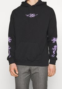 Felpa nera con design tribale viola sul petto e grafiche di drago su entrambe le maniche, indossata con pantaloni grigi a scacchi.