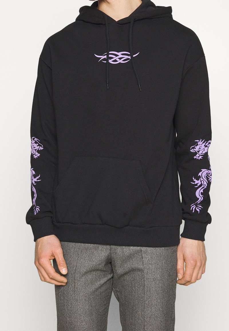 Felpa nera con design tribale viola sul petto e grafiche di drago su entrambe le maniche, indossata con pantaloni grigi a scacchi.
