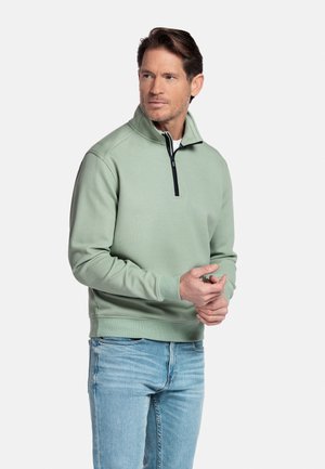 Mann trägt hellgrünen, strukturierten Pullover mit Reißverschluss am Kragen und helle blaue Jeans, steht mit verschränkten Händen und schaut zur Seite.