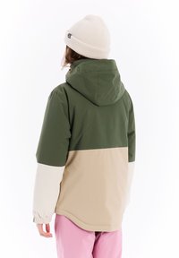 Veste à capuche avec une partie supérieure verte, un milieu beige et une partie inférieure blanche. Dispose de poignets élastiques et d'une coupe décontractée. Accessoire : bonnet en laine.