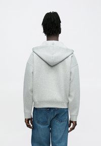 Γκρι φούτερ με κουκούλα και τσέπη καγκουρό, κατασκευασμένο από λείο ύφασμα. Φορεμένο με φαρδιά μπλε τζιν, δείχνοντας άνετη εφαρμογή.