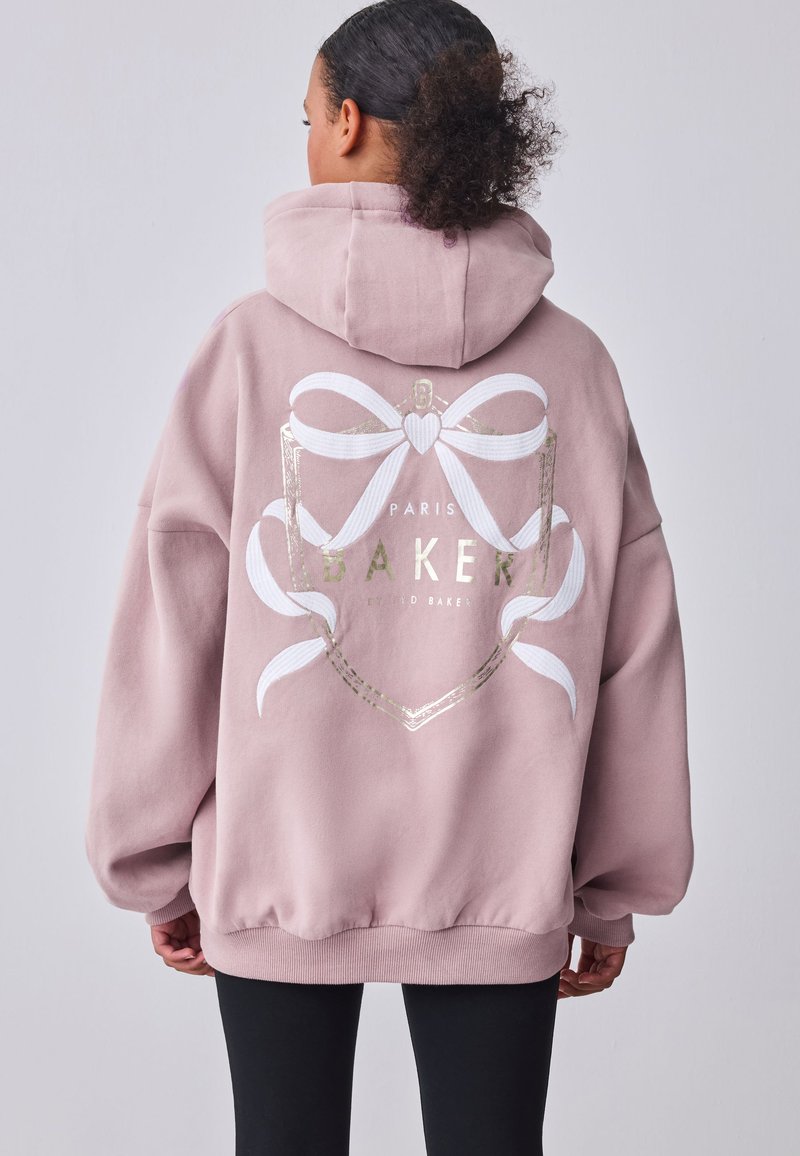 Frau trägt übergroßen staubrosa Hoodie mit weißer Schleife und „Paris Baker“-Schriftzug auf dem Rücken, kombiniert mit schwarzen Hosen.