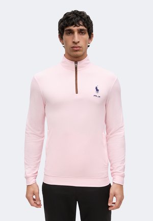 Junger Mann trägt einen hellrosa langärmeligen Pullover mit Reißverschluss und kleinem dunkelblauem Polo-Logo auf der linken Brust.