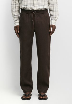 Uomo che indossa pantaloni marrone scuro con coulisse, sandali infradito neri e una camicia bianca a quadri con maniche lunghe e bottoni.
