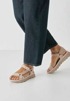Sandales de sport beiges avec des brides texturées et des fermetures réglables ; semelle épaisse, rainurée et de couleur claire ; portées avec un pantalon en jean foncé.