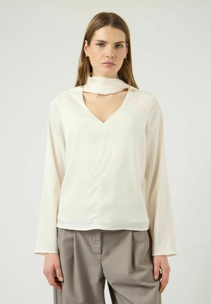 Mlada ženska, oblečena v belo majico z dolgimi rokavi in izrezom v obliki črke V ter bež ohlapne hlače, stoji pred enostavnim svetlim ozadjem.