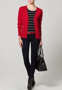 Cardigan rosso su una maglietta a righe bianche e nere, jeans skinny blu, stivali neri alla caviglia e una borsa nera. Collana con catena dorata come accessorio.