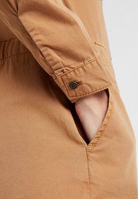 Main vêtue d'une veste beige avec poignet boutonné, doigts partiellement dans la poche latérale d'un vêtement en tissu beige assorti.