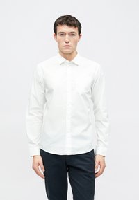 Chemise blanche à manches longues avec col et une poche poitrine unique, fabriquée en tissu lisse. Associée à un pantalon foncé.