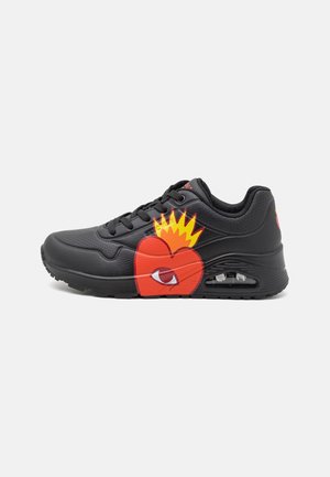 Zapatilla negra con un corazón rojo y un gráfico de llama amarilla, con una suela visible con amortiguación de aire y cordones negros.