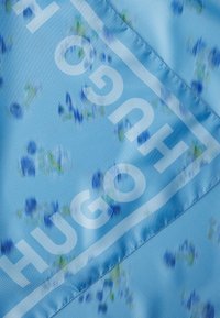 Lichtblauwe stof met een bloemendessin in blauw en groenaccenten. Grote witte tekst leest "HUGO" diagonaal over het ontwerp.