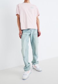 Camiseta de manga corta rosa claro con un pequeño logotipo azul, combinada con jeans azul claro y zapatillas blancas con acentos morados y naranjas.