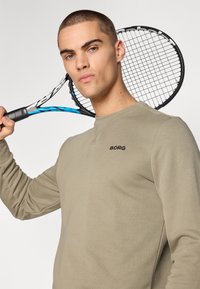 Long-sleeve, olive green sweatshirt with crew neckline and black "BORG" logo on the chest. Model holds a tennis racquet over the shoulder.