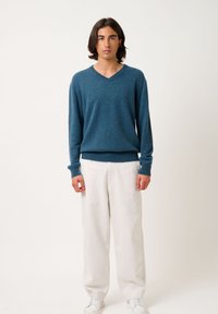 Tealfarbener V-Ausschnitt-Strickpullover aus weichem Material, getragen mit weißen Leinenhosen. Das Model steht vor einem neutralen Hintergrund.