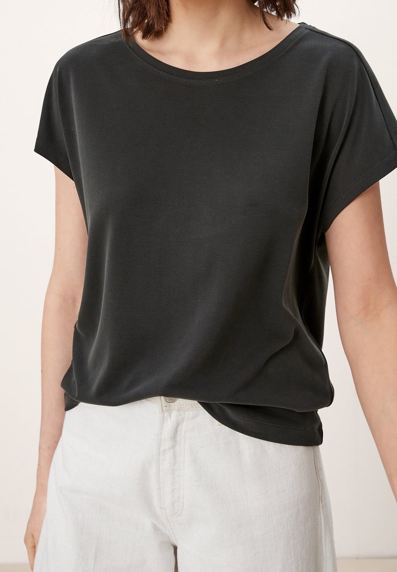Femme portant un haut noir ample à manches courtes et un pantalon blanc taille haute sur un fond uni.