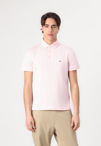 Camisa polo rosa claro con tapeta de botones, mangas cortas y un pequeño logotipo en el lado izquierdo del pecho. Textura de tela de algodón suave.