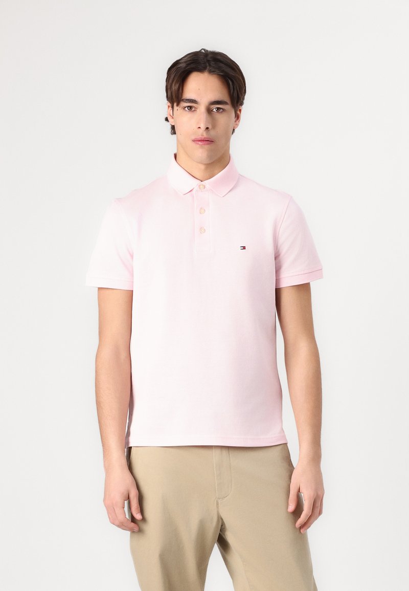 Camisa polo rosa claro con tapeta de botones, mangas cortas y un pequeño logotipo en el lado izquierdo del pecho. Textura de tela de algodón suave.