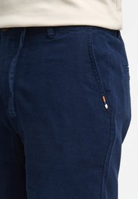 Navy cotton pants with a straight leg design, featuring a visible drawstring and a small multi-colored fabric label on the side pocket.
