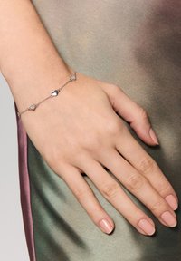 Srebrna bransoletka łańcuszkowa z naprzemiennie ułożonymi zawieszkami w kształcie serca oraz małymi, błyszczącymi detalami. Dłoń spoczywa na delikatnie zabarwionej, teksturowanej tkaninie.