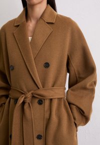 Manteau en laine marron à double boutonnage avec boutons noirs et ceinture nouée, porté sur un haut blanc, les mains dans les poches.